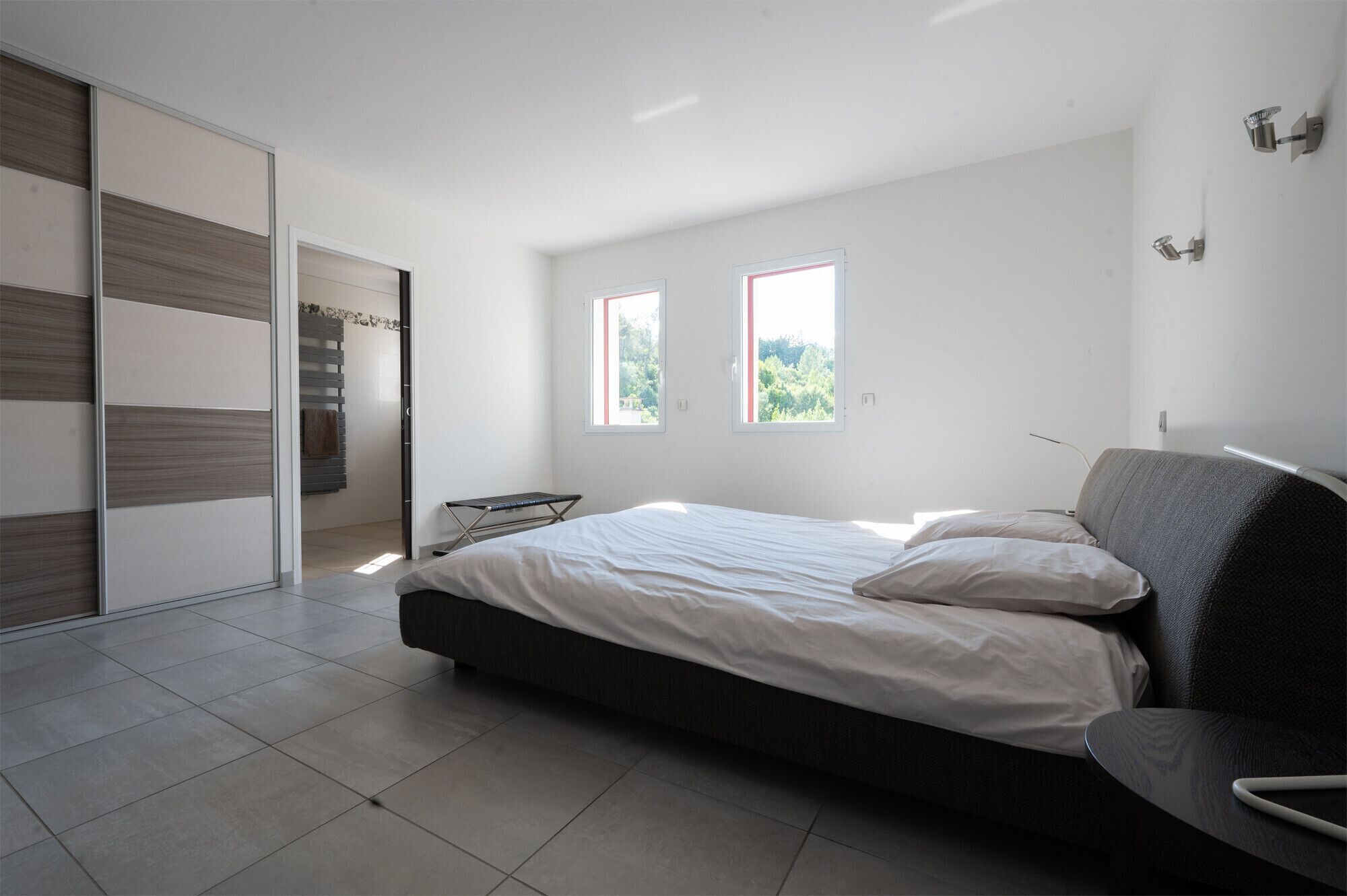 A modern bedroom with a large bed, a bench, and two windows providing natural light.