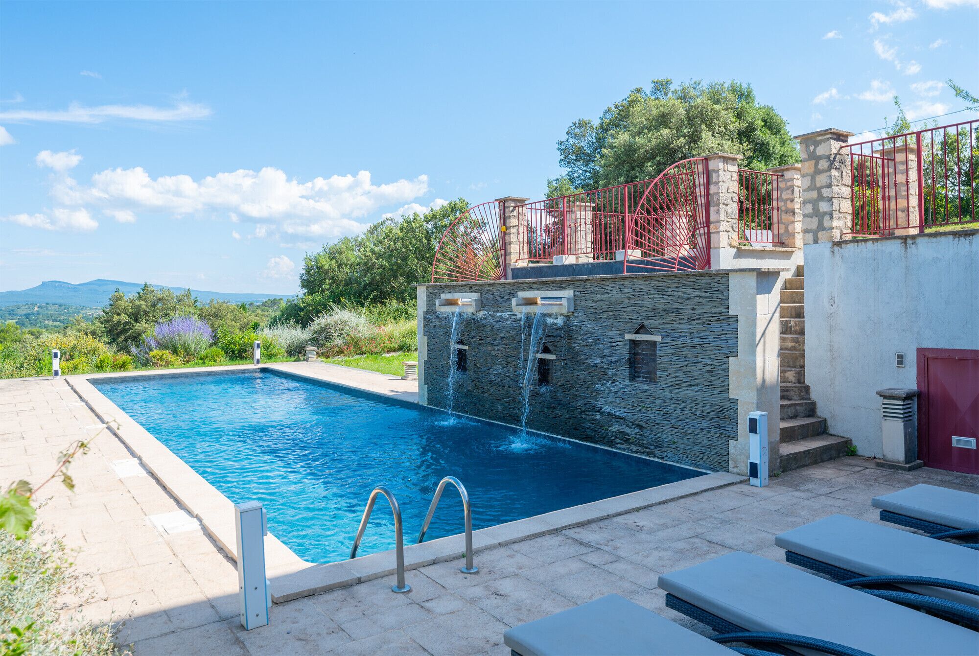 Ein Schwimmbecken mit Steinmauern, einer roten Metalltreppe und Blick auf die Landschaft.