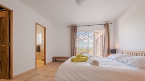Een slaapkamer met een bed, een bankje, een raam met gordijnen en uitzicht naar buiten.