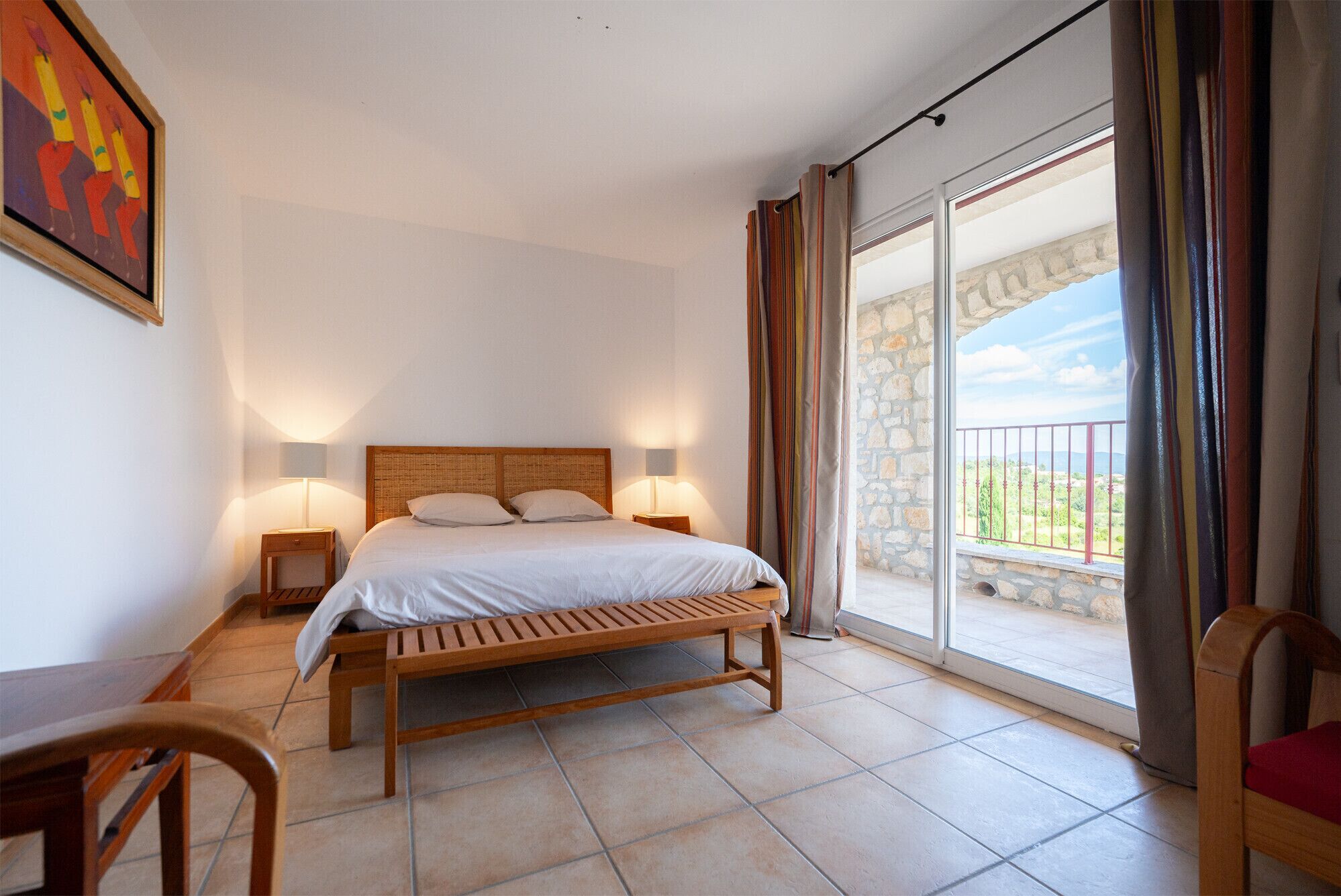 Een slaapkamer met een bed, nachtkastjes, een bankje en uitzicht op een balkon.