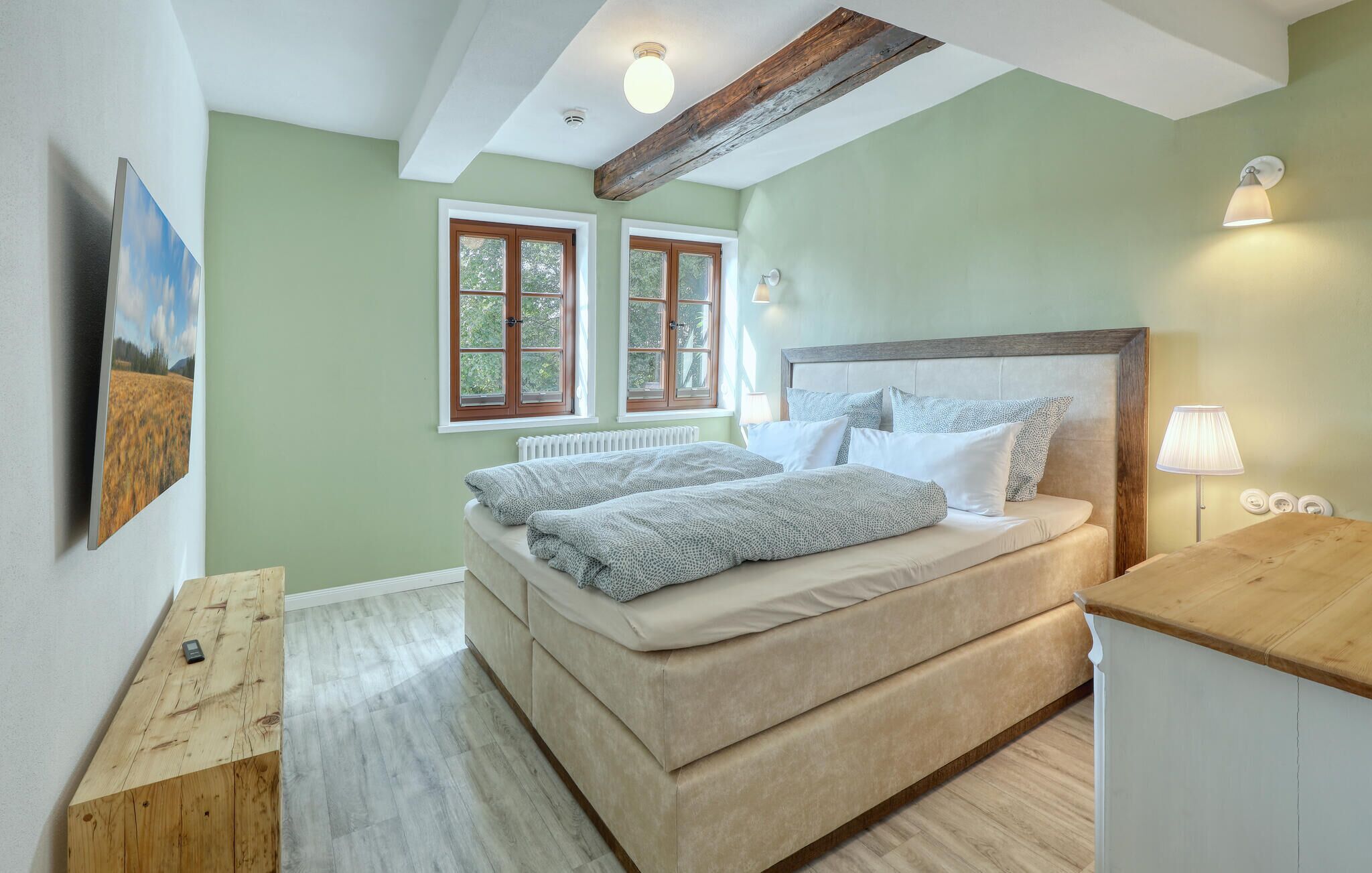 Un dormitorio con cama, dos ventanas, cabecera de madera, lámpara de noche y mesita de noche de madera.