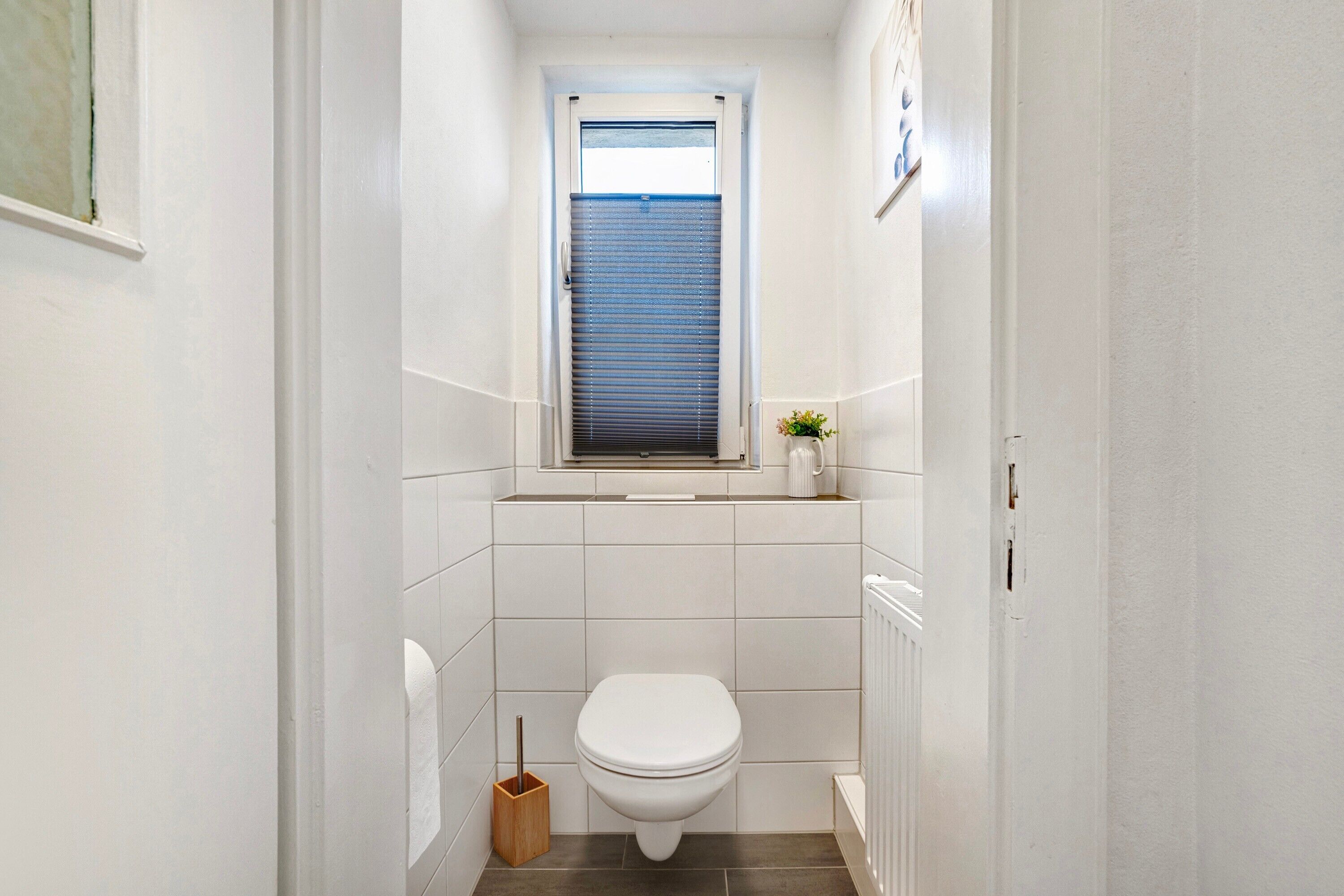 A narrow bathroom with white tiles, a white toilet, a window with blinds, and a small potted plant on the windowsill.