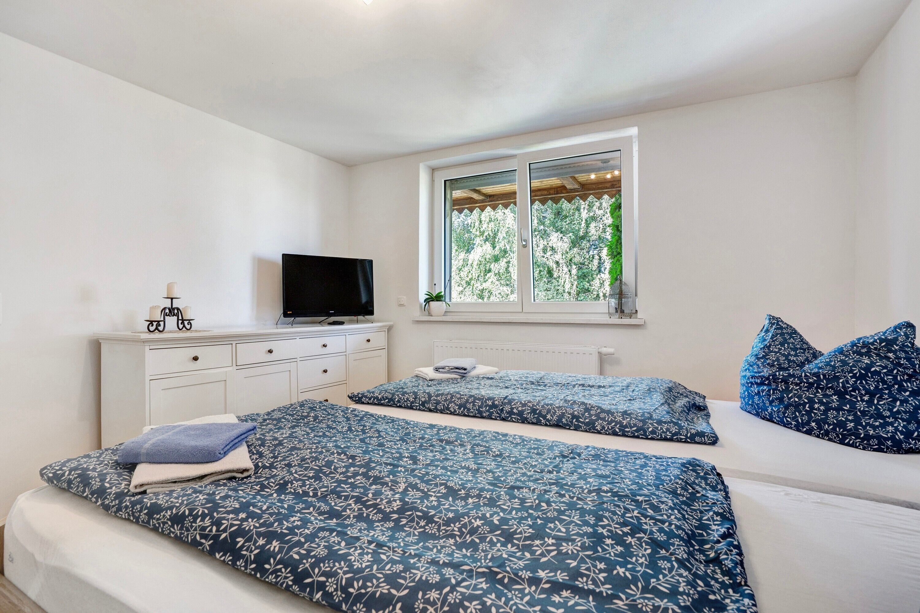 Un dormitorio con dos camas, un televisor sobre una cómoda, una ventana con vista a árboles y un techo blanco.
