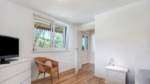 Una habitación con televisión, una silla de mimbre, un aparador blanco, una mesita de noche y una puerta que da a otra habitación.