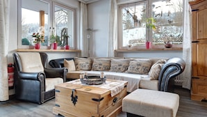 A cozy living room with a sofa, armchairs, a wooden chest, and a window with potted plants. - 'Doppelzimmer Ohne Balkon 26' with Shared Garden and Wi-Fi (St. Georgen)