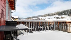 A snow-covered patio with a table and chairs, a wooden fence, and a view of a snowy landscape and houses in the distance.