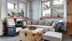 A cozy living room with a sofa, armchairs, a wooden chest, and a window with potted plants.