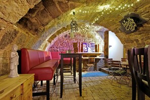 Un intérieur aux murs de pierre, avec une table en bois, des sièges rouges et des guirlandes lumineuses.