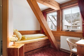 A small, cozy room with a wooden bed, a table, a chair, and a window with a view of a snowy landscape.