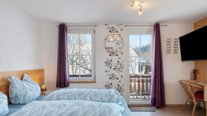 A bedroom with a bed, a desk, a chair, a TV, and a balcony with curtains.