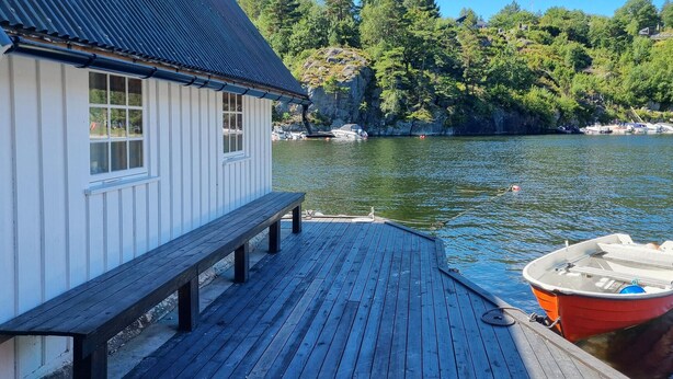 A wooden dock with a bench, a small white building with windows, and a red boat docked at the end.