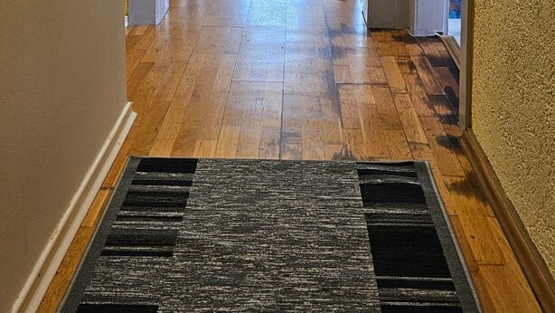 Un couloir doté d’un parquet en bois, d’un tapis noir et d’une porte qui donne sur une pièce lumineuse.