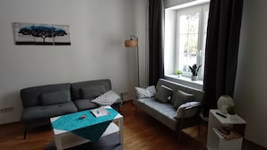 Un salon avec un canapé gris, une petite table recouverte d’une nappe turquoise, un lampadaire et une fenêtre avec des rideaux.