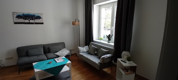 Un salon avec un canapé gris, une petite table recouverte d’une nappe turquoise, un lampadaire et une fenêtre avec des rideaux.