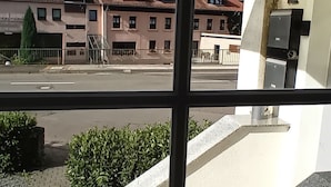 Une vue par la fenêtre sur un immeuble avec un escalier menant à une zone abritée, un panneau indiquant le numéro « 12 », et une rue bordée de maisons en arrière-plan.