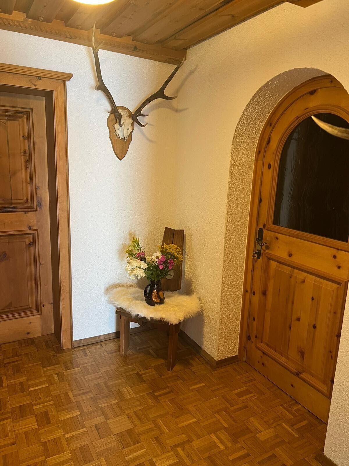 A wooden door, a wall-mounted deer skull, a small table with a vase of flowers, and a wooden bench.