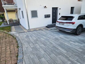 Un coche blanco aparcado en una entrada de casa con una vivienda moderna, un balcón y un pequeño jardín.