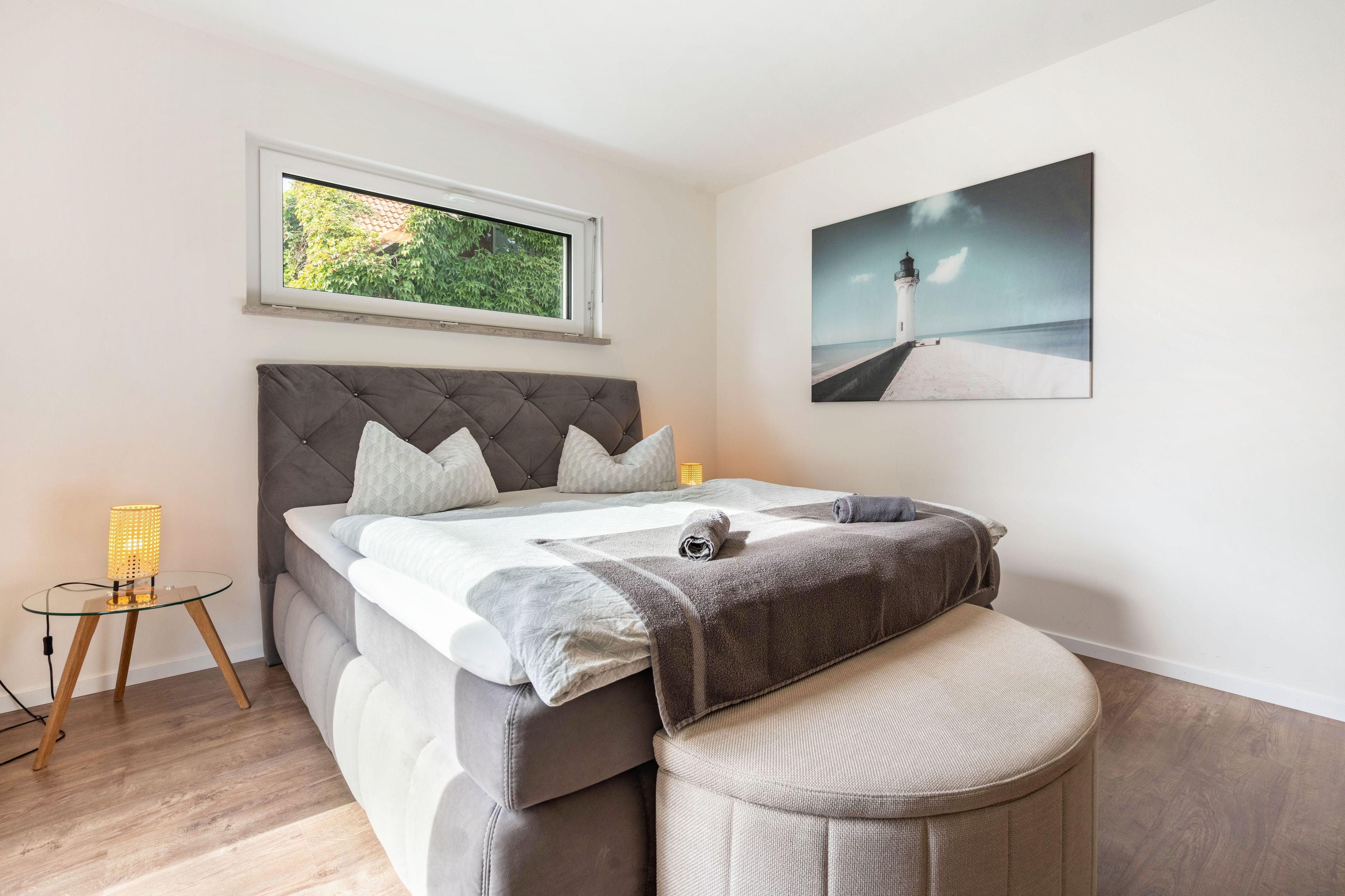 Ein Schlafzimmer mit einem Bett, einem Fenster, einem Nachttisch, einer Lampe und einem Bild an der Wand.