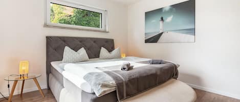 A bedroom with a bed, a window, a bedside table, a lamp, and a painting on the wall.
