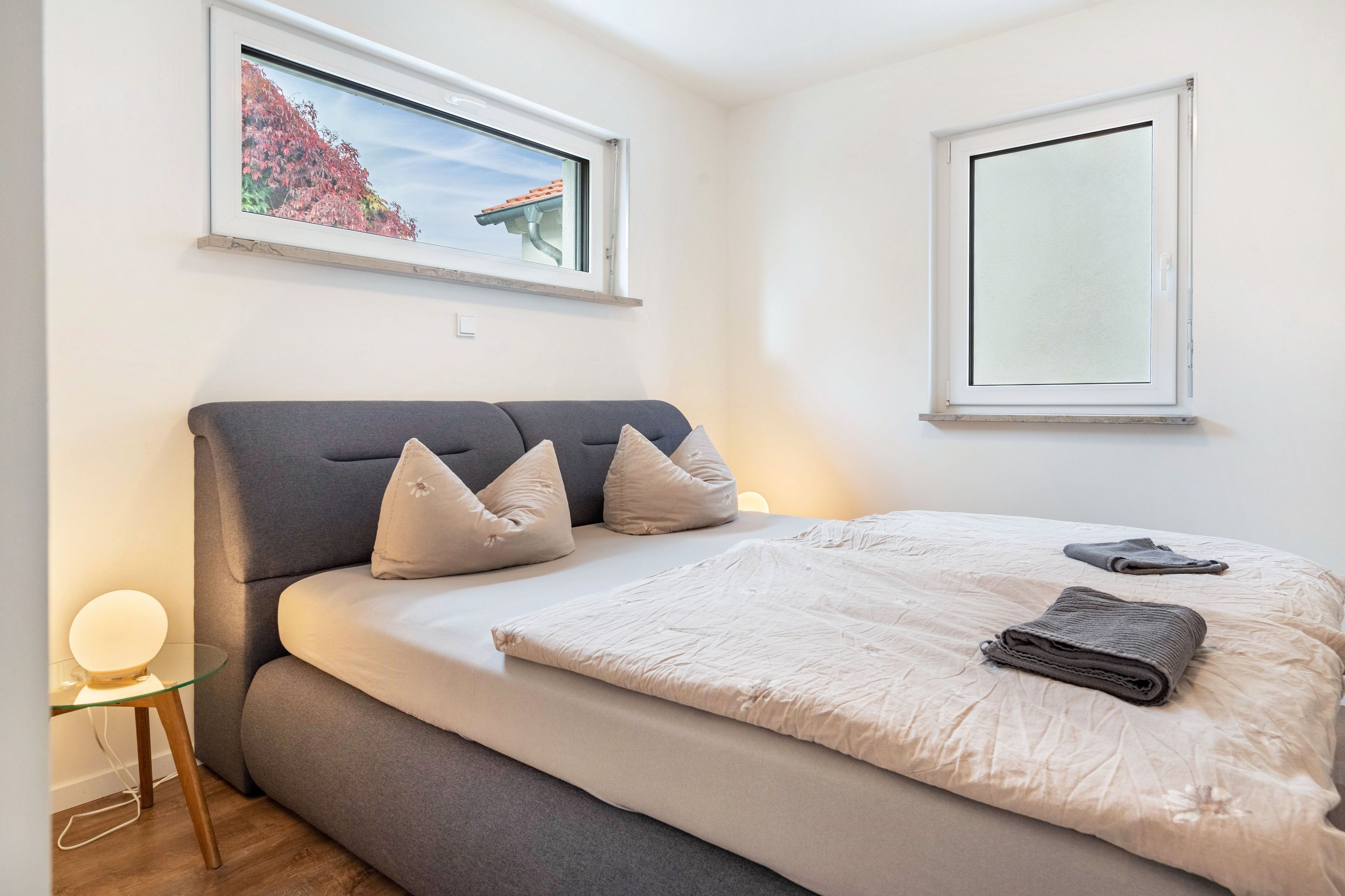 Ein Schlafzimmer mit einem Bett, Kissen, einem Nachttisch mit Lampe und einem Fenster mit Blick auf Bäume.