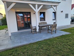 Eine überdachte Terrasse mit Tisch und Stühlen, eine Glastür und ein kleines Fenster.