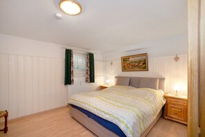 Ein Schlafzimmer mit einem Bett, einem Nachttisch, einer Lampe, einem Bild an der Wand und einem Fenster mit Vorhängen.
