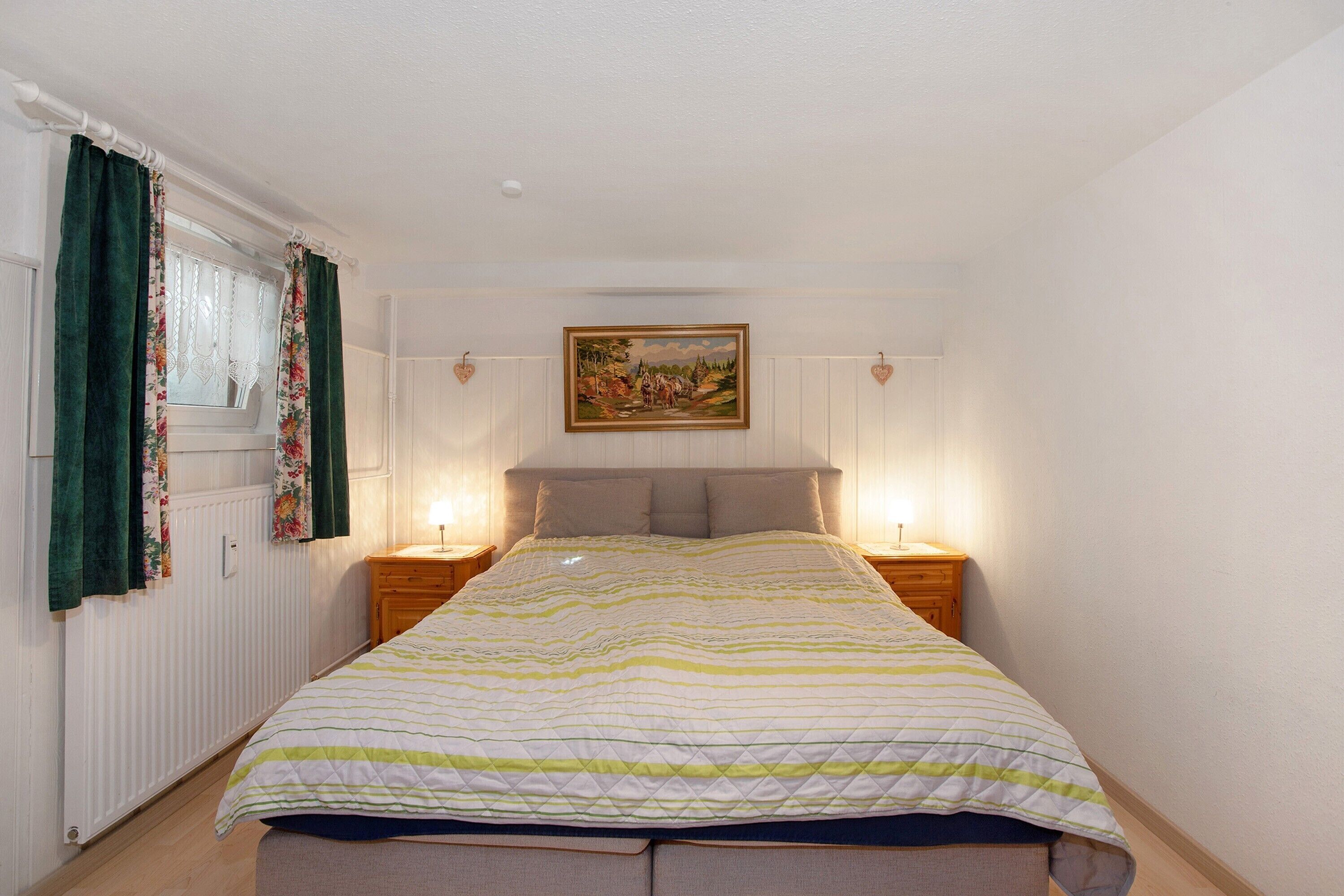Een slaapkamer met een bed, twee nachtkastjes, een raam met gordijnen, een aan de muur hangend schilderij en een radiator.