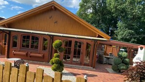 A wooden building with a covered patio area, large windows, and a fenced garden. - Holiday Home 'Idylle Ferienhaus Ems' with Lake View, Wi-Fi and Air Conditioning (Walchum)