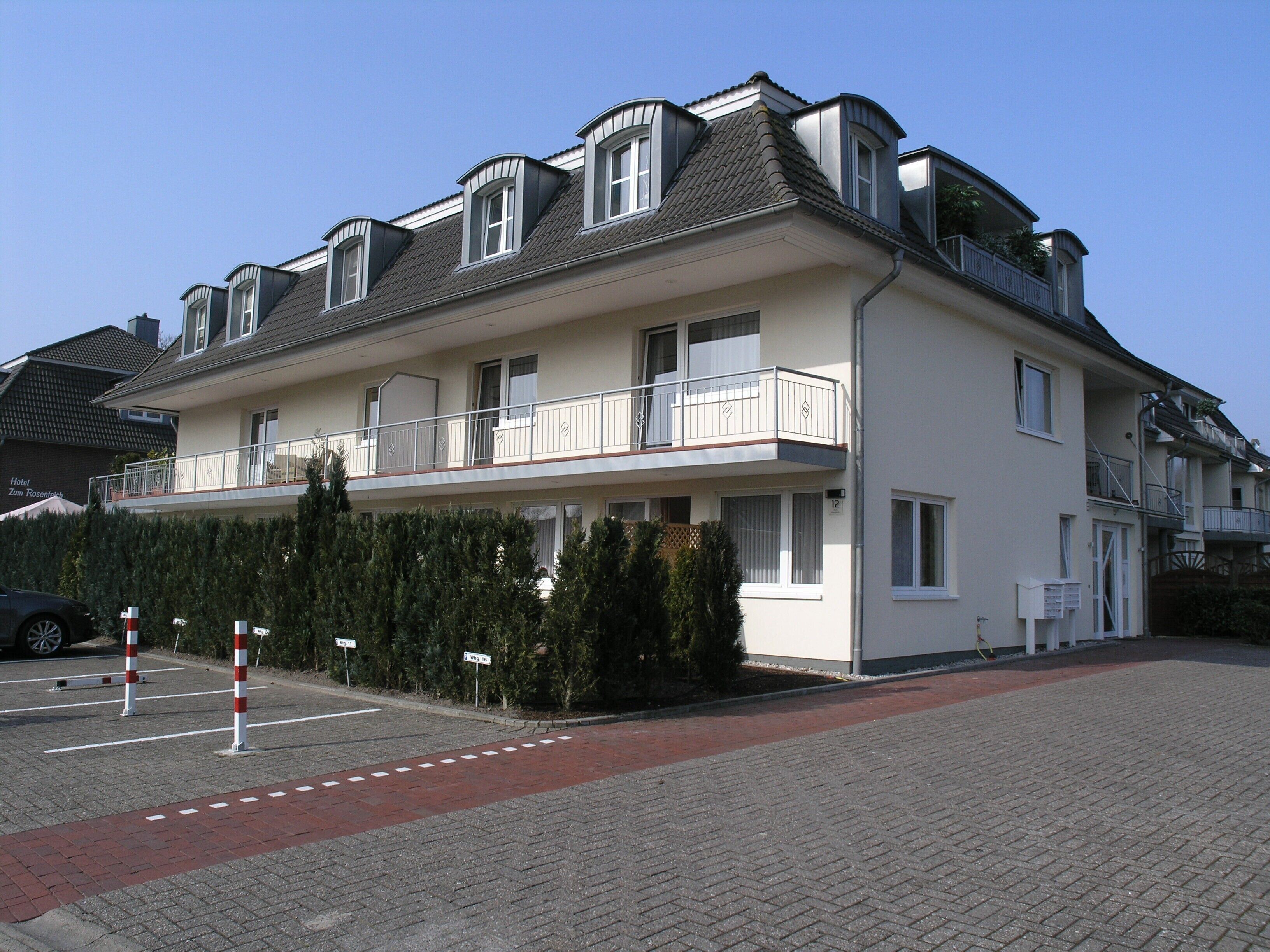 Un hôtel à plusieurs étages doté d’un balcon au deuxième étage, d’un parking avec des places délimitées et d’une haie à l’avant.