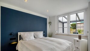 Un dormitorio con cama, una lámpara de pared, una ventana con vistas y un pequeño armario blanco.