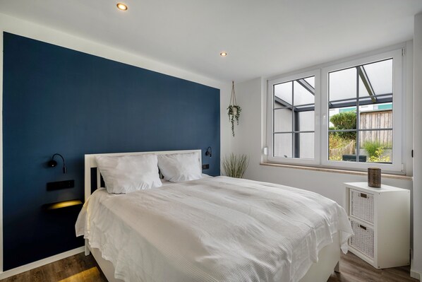 Une chambre à coucher avec un lit, une lampe fixée au mur, une fenêtre donnant sur un beau paysage et une petite armoire blanche.
