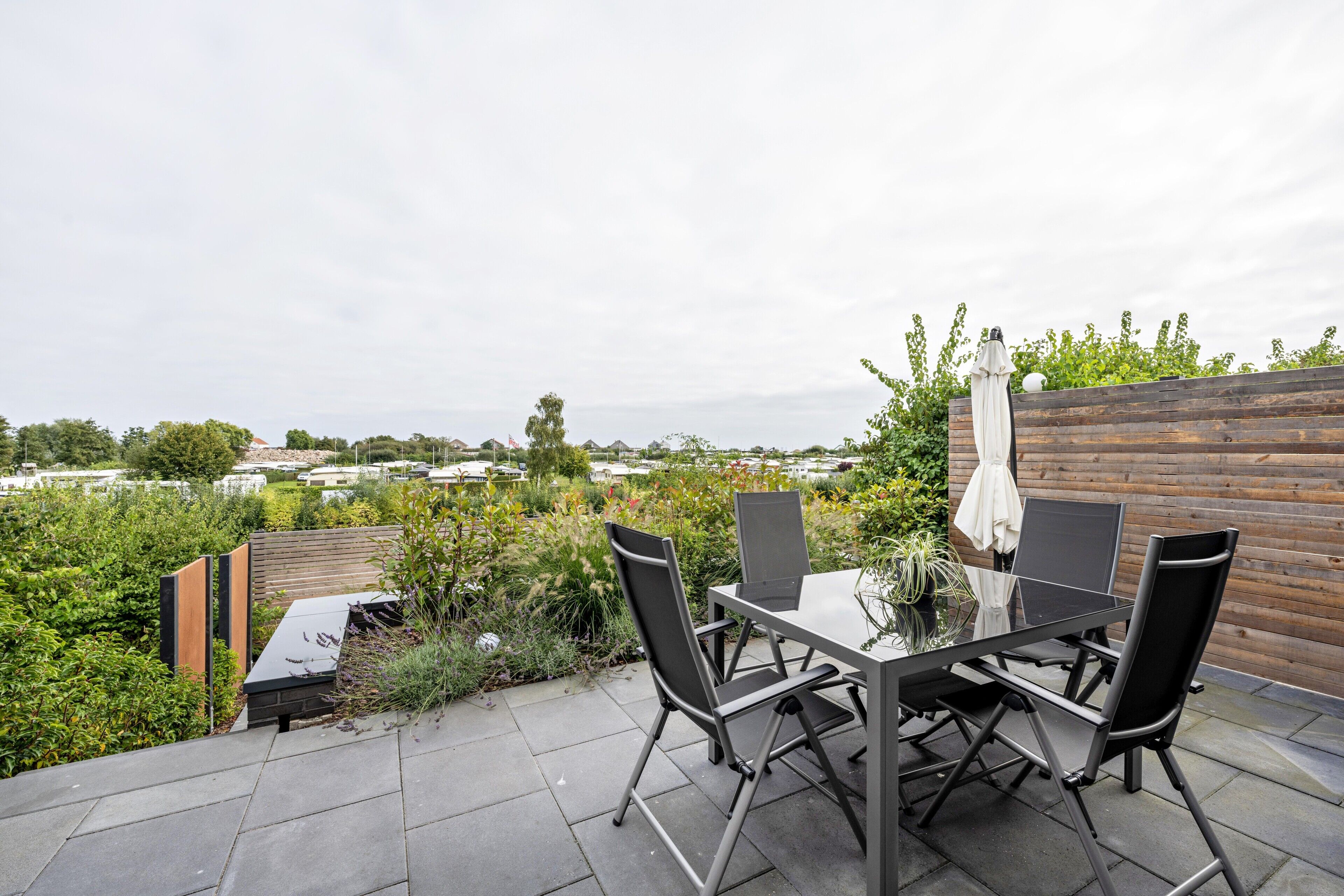 En udendørs terrasse med et bord og stole, omgivet af grøn vegetation og et træhegn.