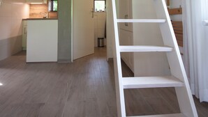Interior em madeira com escada branca que leva a um mezanino, cozinha com janela e banheiro com chuveiro.
