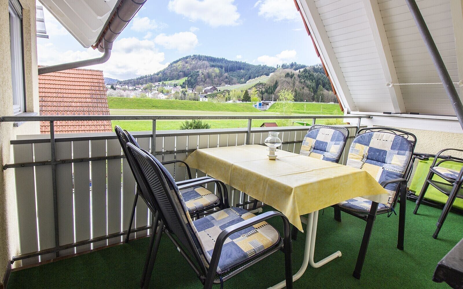 A balcony with a table and chairs, a view of a green landscape, and a building in the background.
