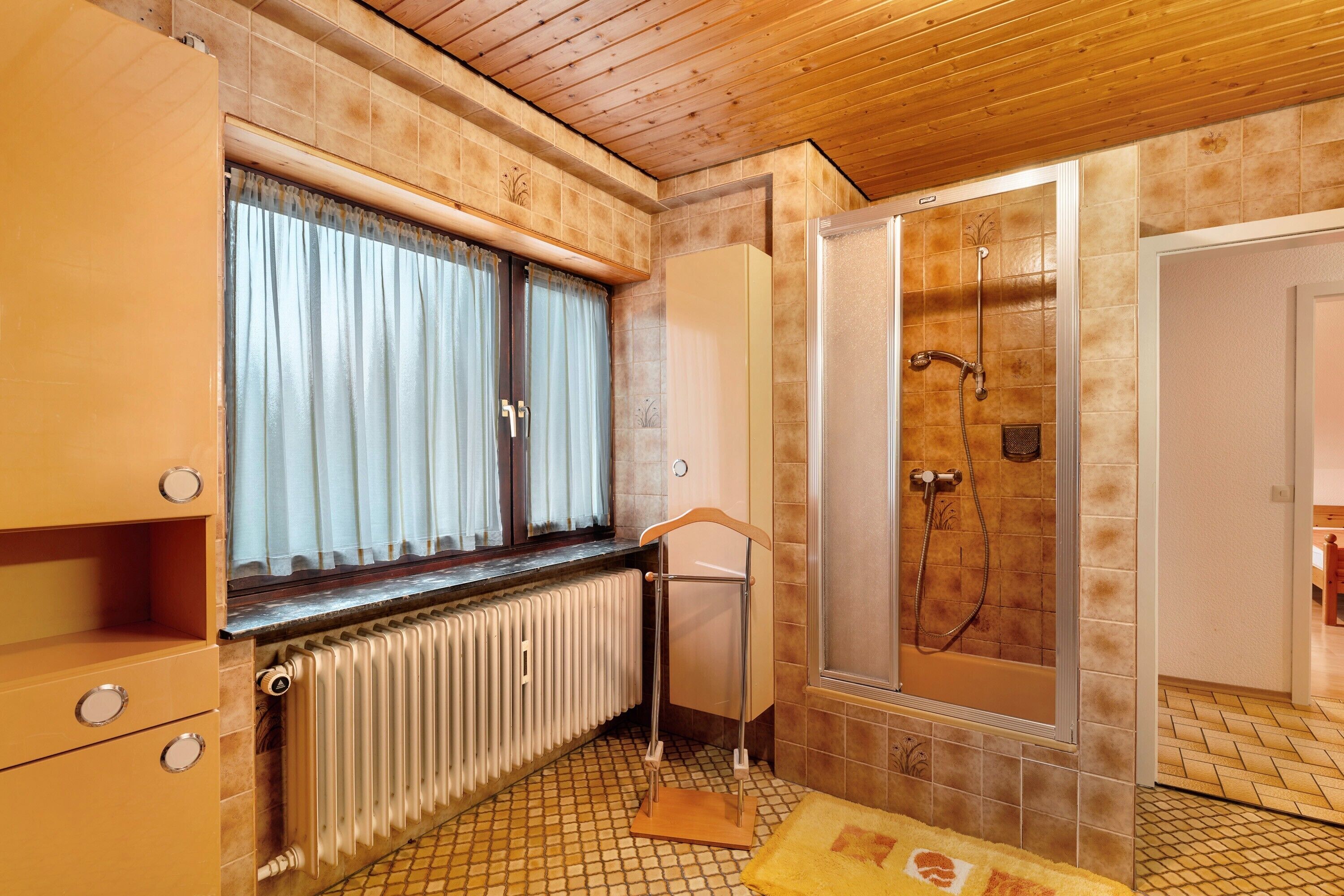Ein Badezimmer mit Dusche, Heizkörper, einem Fenster mit Vorhängen und Holzschränken.