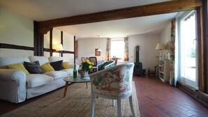 Un salón con un sofá blanco, un sillón estampado, una mesita con un florero, una chimenea y una ventana con cortinas.