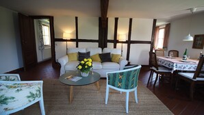 Una sala de estar con un sofá blanco, una mesa redonda con un florero, un sillón estampado y una zona de comedor con una mesa puesta para dos.