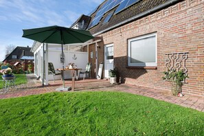 Une terrasse avec une table et des chaises, un parasol vert et un mur en briques équipé de panneaux solaires.