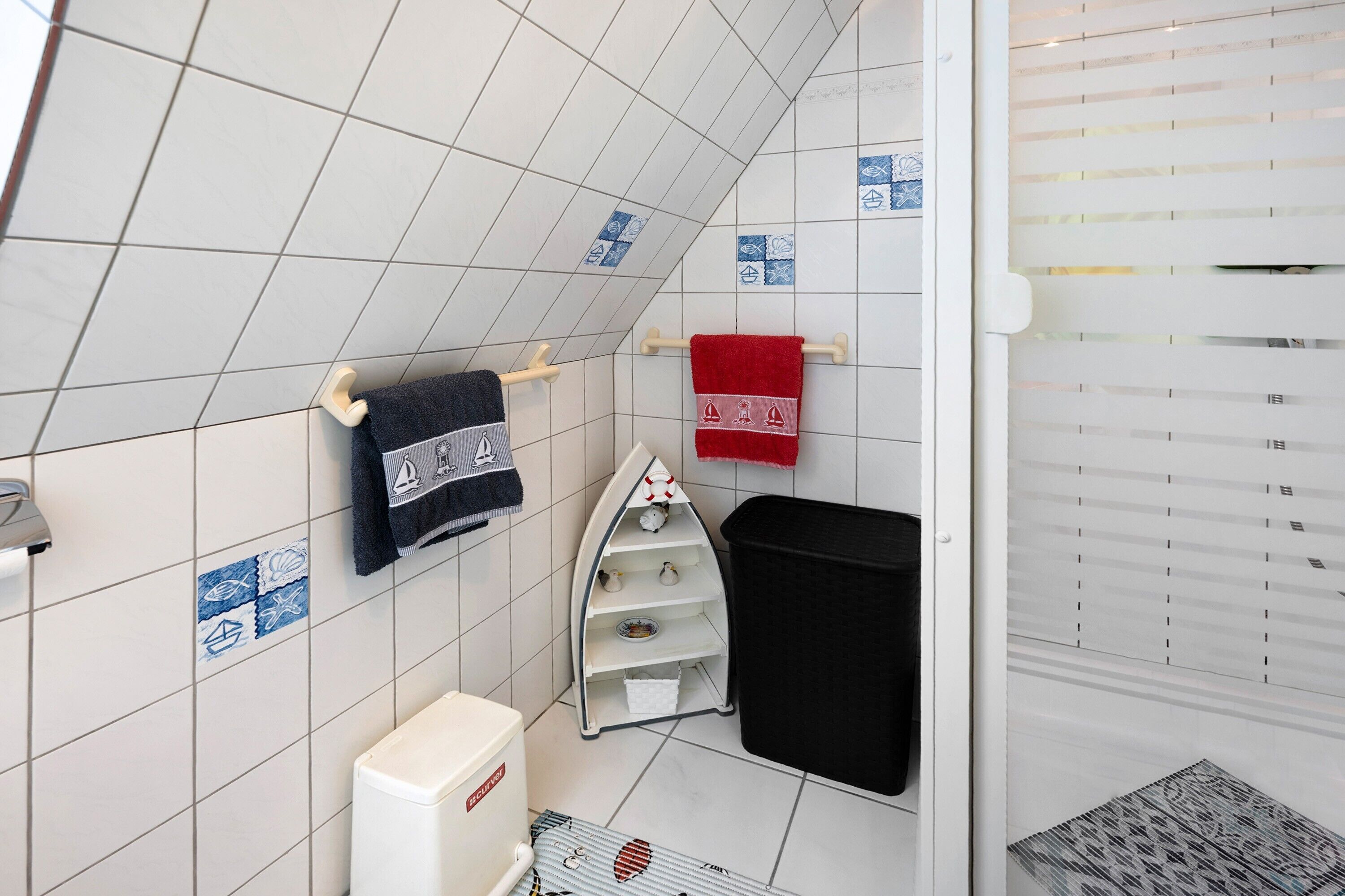 Un bagno con pareti piastrellate di bianco, un water bianco, un cestino nero e un portascopino con due asciugamani.