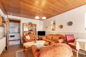 Ein gemütliches Wohnzimmer mit Holzbalkendecke, einem Fernseher, einem Sofa, einem roten Sessel und einem Esstisch.