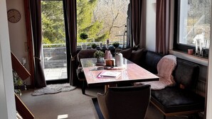 Un salon moderne avec une table à manger, un canapé noir et une vue sur les arbres à travers de grandes fenêtres.