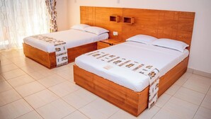 Two wooden beds with white bedding and a patterned runner, a wooden headboard, and a small bedside table with a lamp. - SERENITY FORT HOTEL (Mariakani)