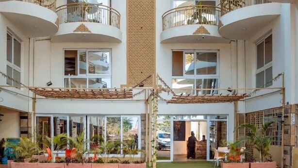 A multi-story hotel with a central atrium, balconies on each floor, and a ground-level entrance with plants and a seating area. - SERENITY FORT HOTEL (Mariakani)