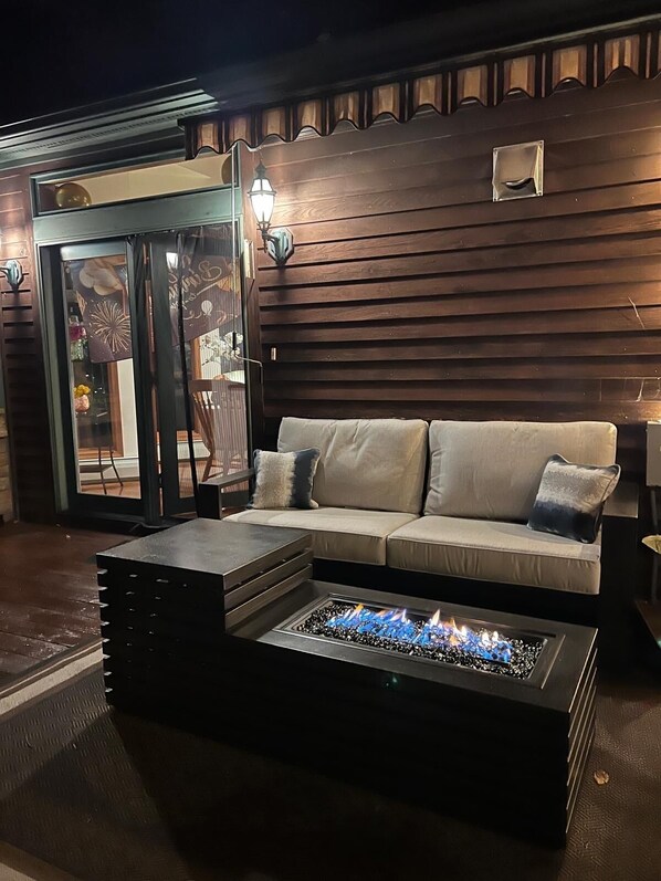 An outdoor seating area with a fire pit table, a cushioned sofa with pillows, and a wooden wall with a window and door.