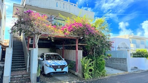 Um edifício residencial com um carro estacionado na entrada, uma escadaria dando acesso à porta e uma área coberta com plantas floridas.