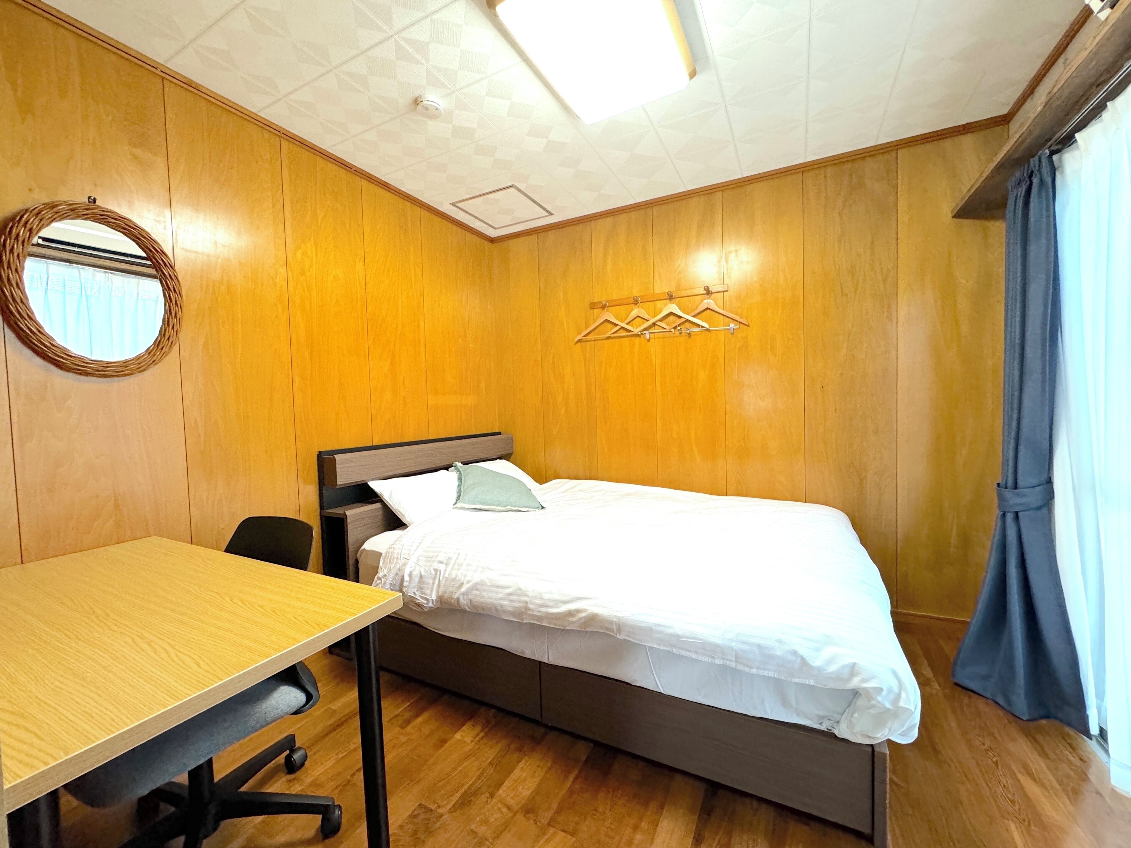 A bedroom with a bed, a desk, and a chair. There is a round mirror on the wall and a clothes rack above the bed.