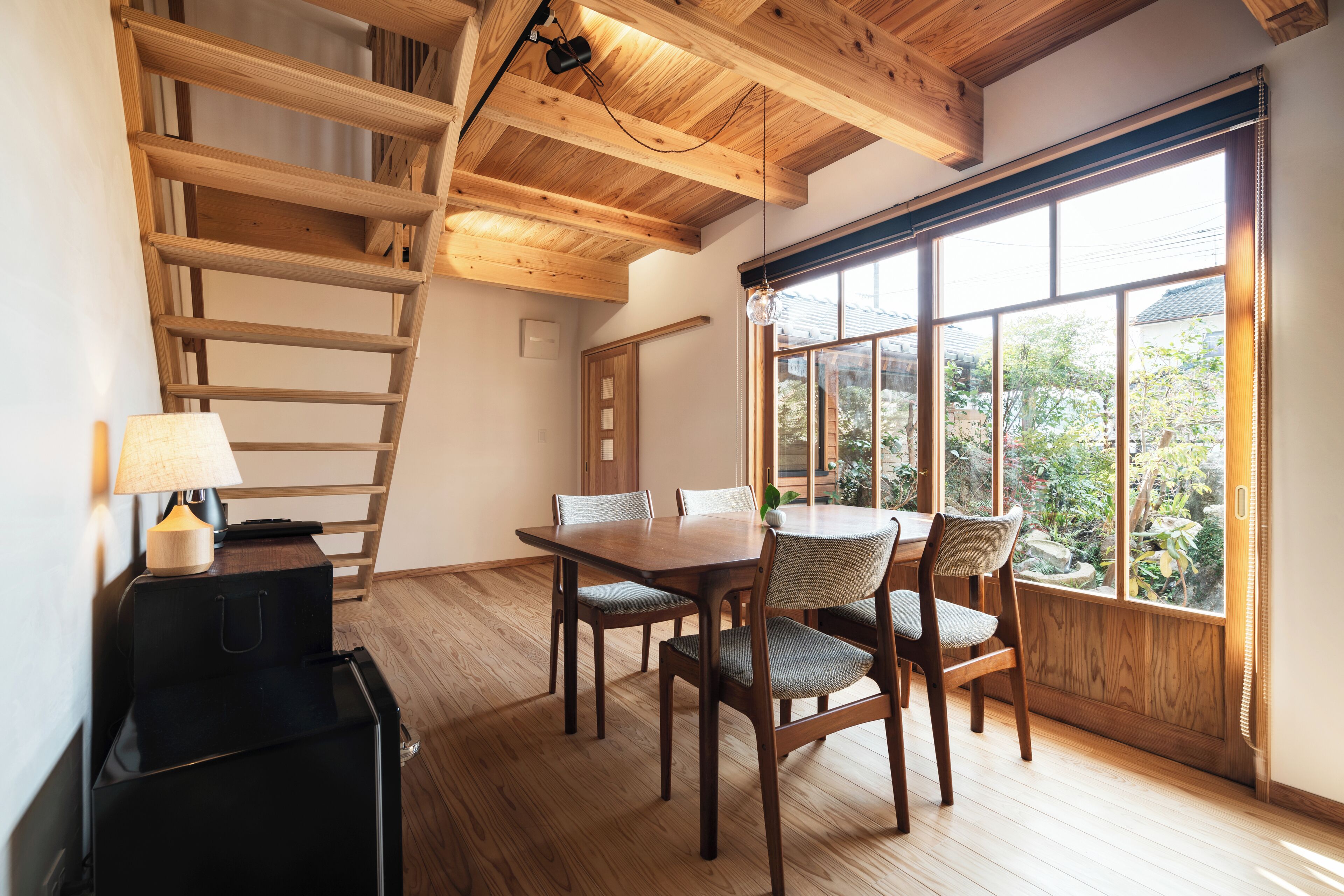 A wooden dining table with chairs, a lamp, and a ladder in a room with large windows and wooden beams.