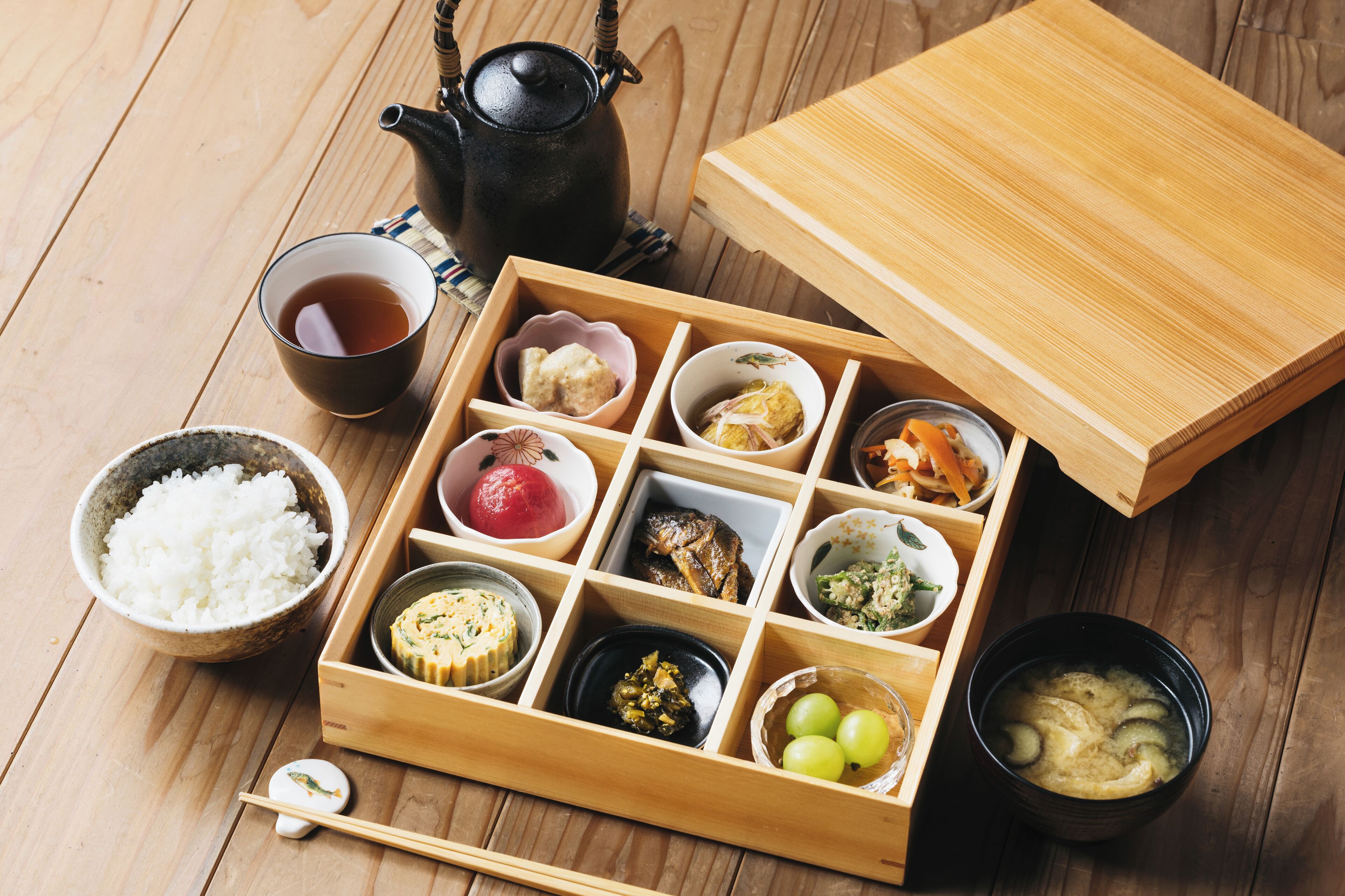 A traditional Japanese bento box with various food items, a teapot, and a cup of tea on a wooden table.