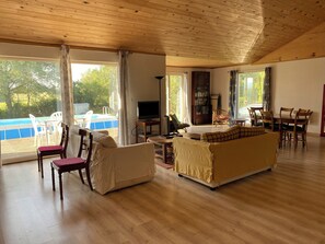 A spacious living room with a wooden ceiling, large windows, a pool, and a dining area.