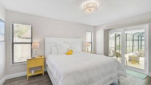A bedroom with a large bed, a nightstand, a window with blinds, and a view of the outdoors through a glass door.
